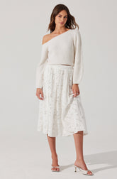 A woman is wearing an off-the-shoulder cream-colored knitted sweater with long sleeves and a loose fit. She pairs it with a white, lace, midi-length skirt that has a soft, flowy silhouette and intricate lace detailing. She completes the outfit with light-colored, open-toe heeled sandals.